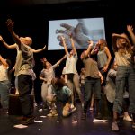 Photo showing group of performers of mixed ages on stage, many holding their arms above their heads with a screen showing a photo of a hand in the background