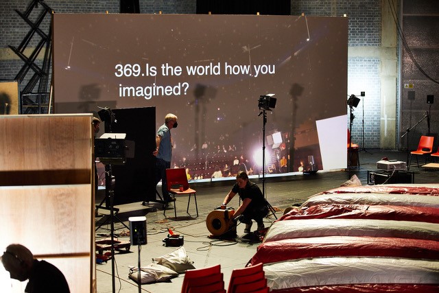 A large board has text that reads: "369. Is the world how you imagined?" In the foreground people are moving cables and working on the stage.