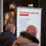 Person wearing a mask stands in a shop window next to poster titled 'This Poem is for You'. They appear to be smiling and reaching out to two people walking past outside.