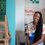 Dalia James, weaver, smiles into camera surrounded by a loom and brightly coloured weavings