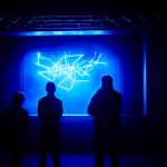 Silhouette of three people looking at a blue neon light artwork