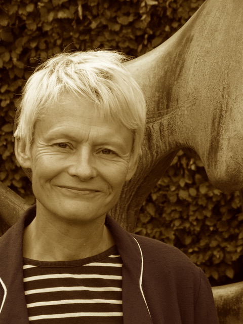 A person pictured in sepia tone colours. They are standing in front of a hedge and wearing a beige jacket.
