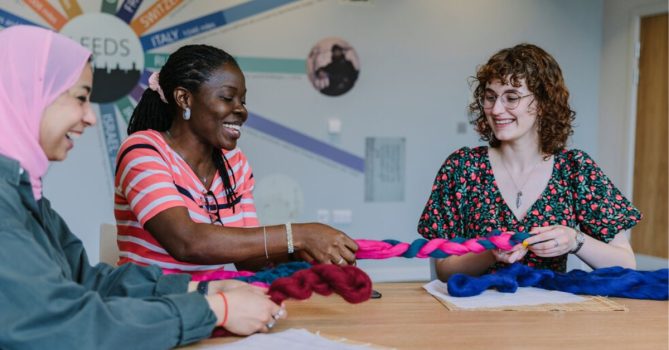 Three young people sat at a table in a gallery setting. They are doing a craft activity twisting together lengths of fabric