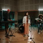 A group of young musicians record in the studio. Three musicians, including a keyboard player, singer, and guitarist, are performing.