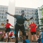A group of people dancing on roller skates. The person at the front has their arms out, is dressed all in black and is wearing knee and wrist pads. The people behind wear red t shirts, one holds a pom pom. in the background are more people, a brick wall and a block of flats.