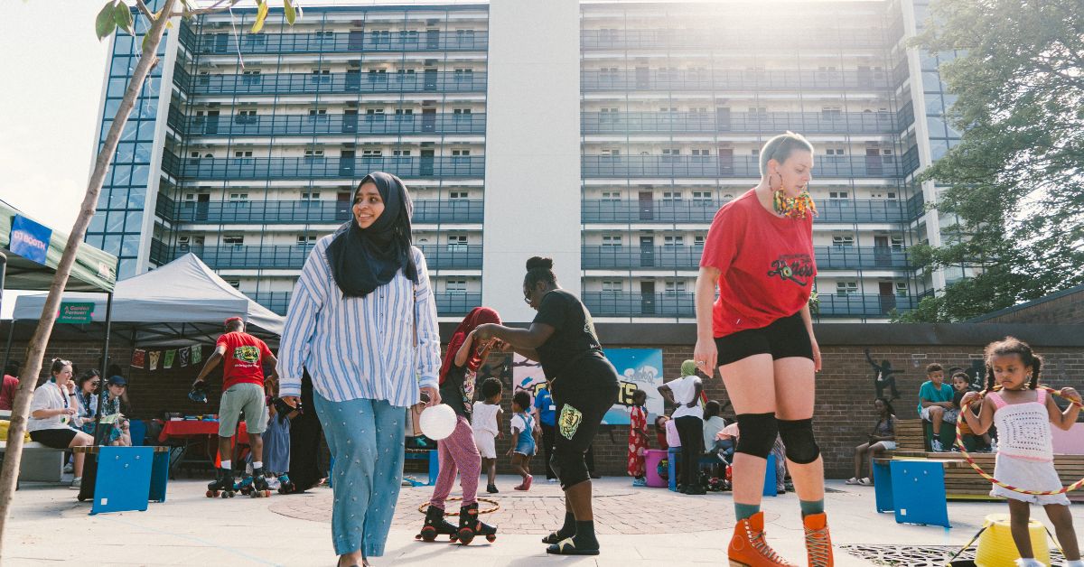 A vibrant community event takes place in front of a block of flats. In the foreground is person wearing a hijab, jeans and a shirt smiling. There is also a person is a red t shirt and roller skates. In the background adults and children are rollerskating and playing.