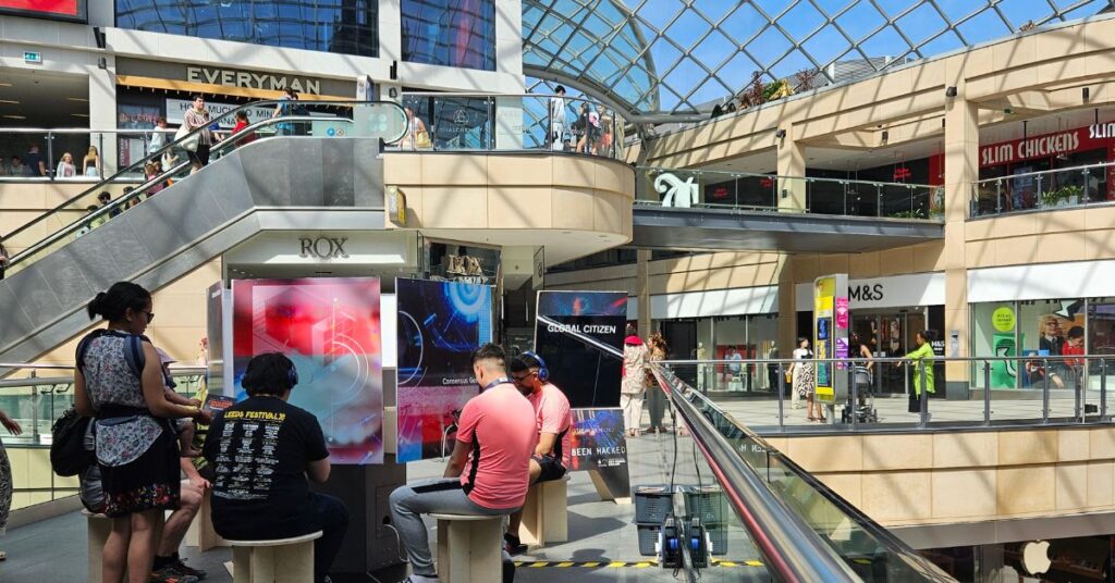 Young people sat on stools in a shopping centre. They have headphones on and are taking part in an immersive experience.