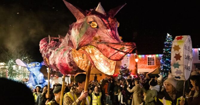 A lantern parade in a town featuring a large, illuminated pink and red dragon lantern with glowing eyes, carried by people in yellow safety vests. A second blue creature lantern with large ears follows behind. 