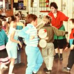Children in a community setting participating in music and dance activities, with some playing tambourines and others dancing. An adult leads the group in a brightly decorated room with wooden floors. This is an archive photo from the 1980s.