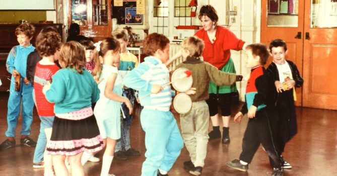 Children in a community setting participating in music and dance activities, with some playing tambourines and others dancing. An adult leads the group in a brightly decorated room with wooden floors. This is an archive photo from the 1980s.