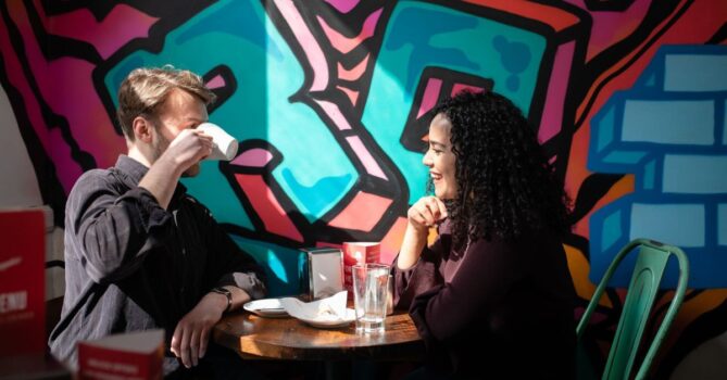 Two people sat opposite each other in a cafe. One is drinking out of white mug. Behind them the wall is decorated in colourful graffiti.
