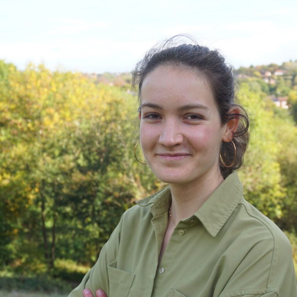 A headshot image of Marion Carneiro. She's wearing a green shirt and is stood in front of trees and greenery.