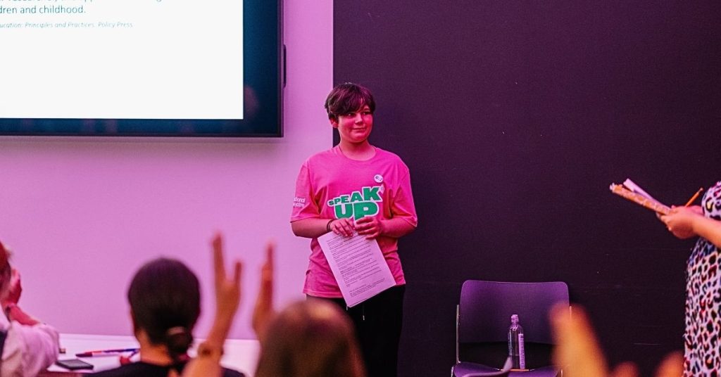 A young person, wearing a pink "speak up" t shirt, presenting to a group of people.