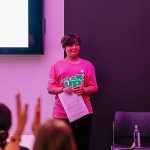 A young person, wearing a pink "speak up" t shirt, presenting to a group of people.