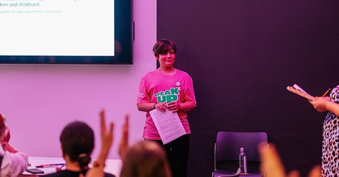 A young person, wearing a pink "speak up" t shirt, presenting to a group of people.