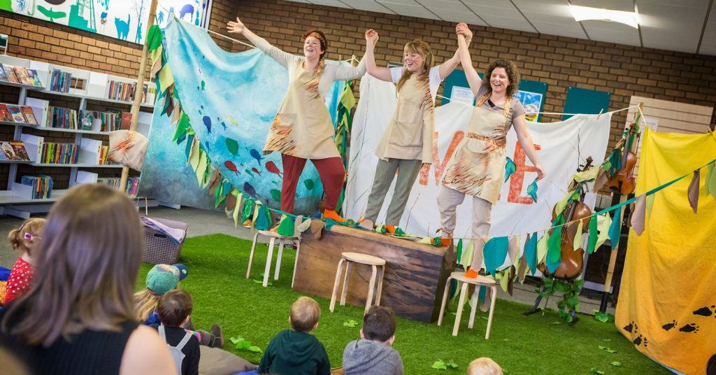 Three performers stood on stools and a chest holding hands with their arms in the air. They are performing a children's show in a library. Children are sat on the floor watching the performance.
