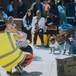 A person in a yellow high vis vest is showing a small group of children a dance. The children are crouched down copying the dancer. Parents sit on blocks watching their children take part.