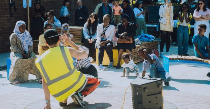 A person in a yellow high vis vest is showing a small group of children a dance. The children are crouched down copying the dancer. Parents sit on blocks watching their children take part.