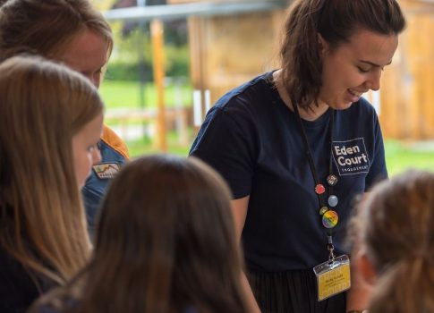 An arts practitioner leading an origami craft activity. Participants are sat around a table by a large window, listening as the leader explains the activity.