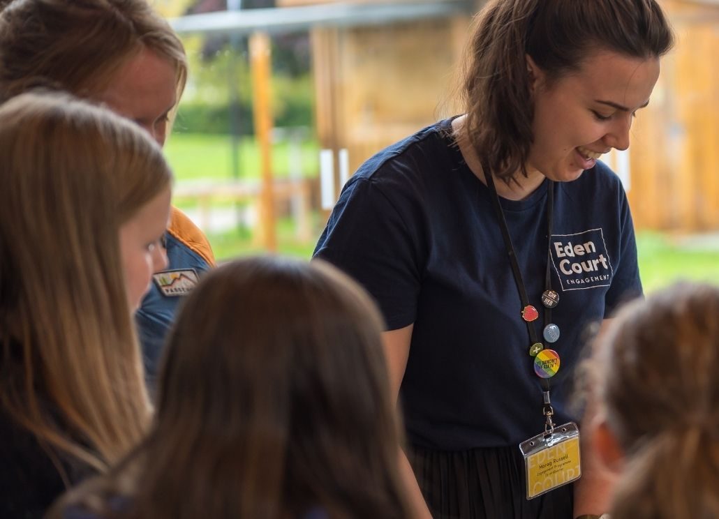 An arts practitioner leading an origami craft activity. Participants are sat around a table by a large window, listening as the leader explains the activity.