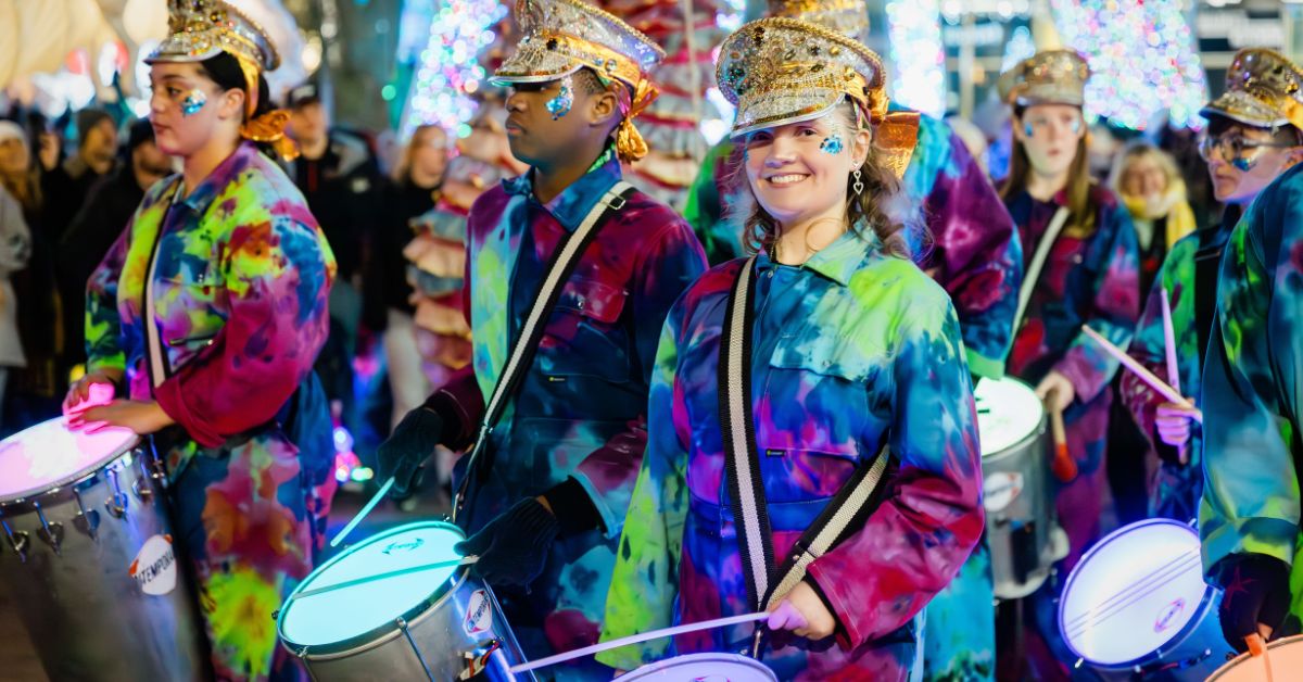 A group of young performers, wearing brightly coloured outfits and fancy hats. They each have a drum and drumsticks.