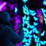 A person in a coat and hat recording a blue light installation on a phone.