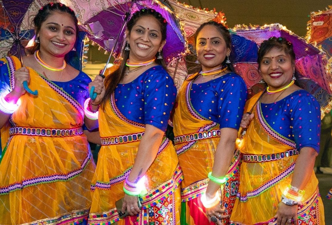 Six women stood together wearing colourful saris and holding parasols. They are each wearing a colourful, glowing wristband
