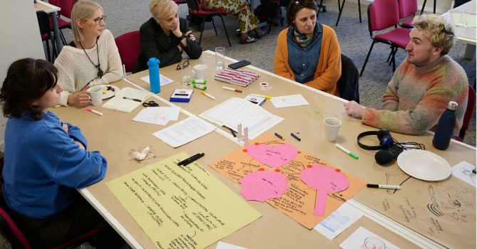 A group of people sat around a table in deep discussion. There's pink and yellow paper, pens and other stationery on the table.