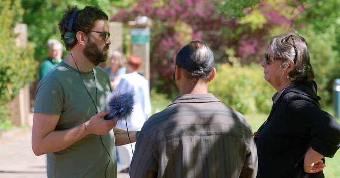 Three people stood talking together outside on a sunny day. One is wearing headphone and holding a microphone.