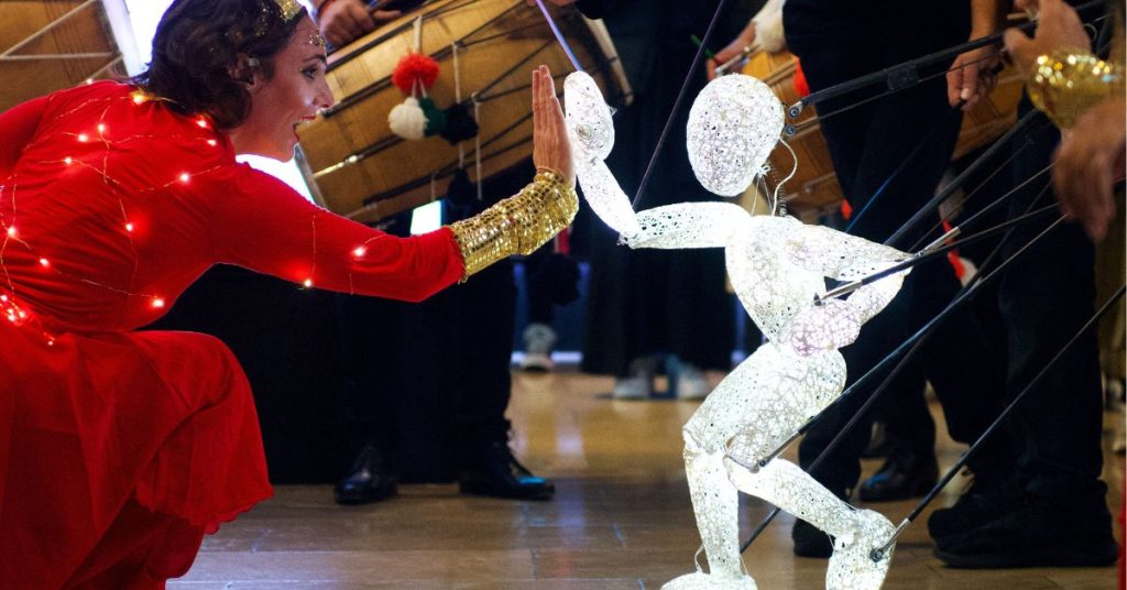A glowing figure of a puppet gives a high five to a person in a red sari.